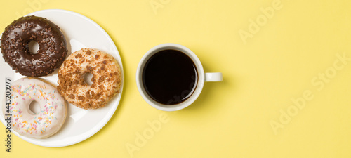 Top view photo of cup of coffee and plate with three different donuts on isolated light yellow background with empty space