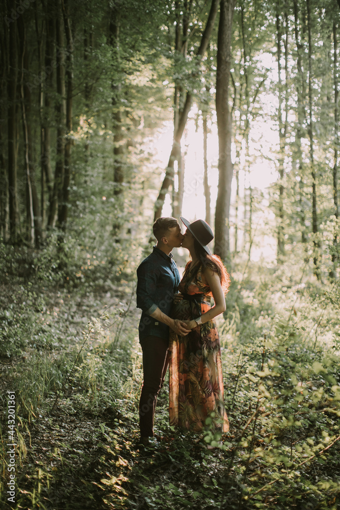 Happy pregnant woman and her man, standing hugging each other, posing on the background of beautiful sunset in the forest among trees, and kissing. Happy pregnancy concept