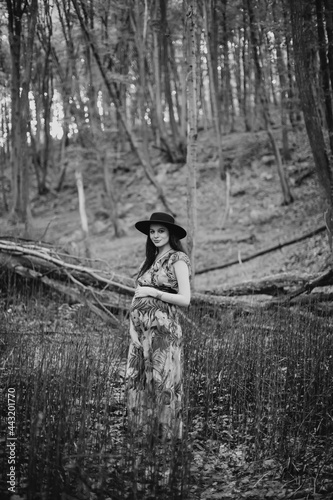 Beautiful young pregnant Caucasian woman, wearing long dress and black stylish hat, relaxing walking in the summer forest