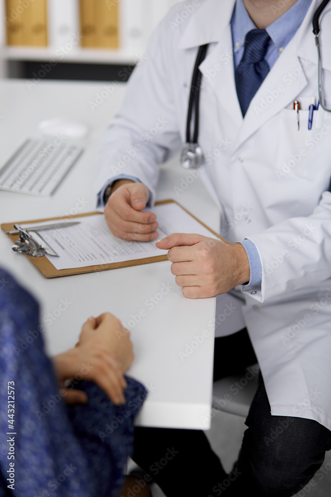 Unknown bearded doctor and patient woman discussing current health examination while sitting in sunny clinic, close-up. Medicine concept Unknown bearded doctor and patient woman discussing current health examination while sitting in sunny clinic, close-up. Medicine concept