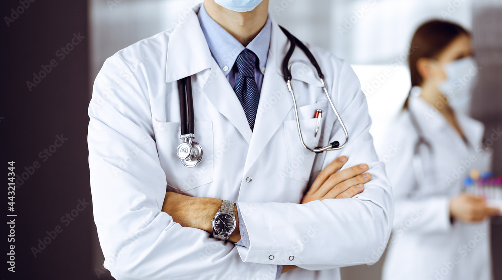 Unknown male doctor in protective mask standing with arms crossed in clinic. Medicine concept during Coronavirus pandemic. Covid 2019 Unknown male doctor in protective mask standing with arms crossed in clinic. Medicine concept during Coronavirus pandemic. Covid 2019