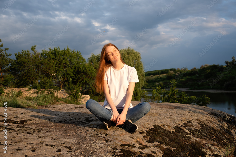 Nature, wellbeing, Relaxation, meditation and mental health. Interacting, Thriving with nature. Calming and enchanting effect. Young redhead woman sitting on rock and watching on river on sunset