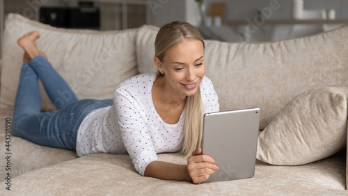 Smiling young woman using tablet, relaxing lying on cozy couch at home, happy attractive female looking at modern device screen, enjoying lazy leisure time with gadget, chatting in social networks