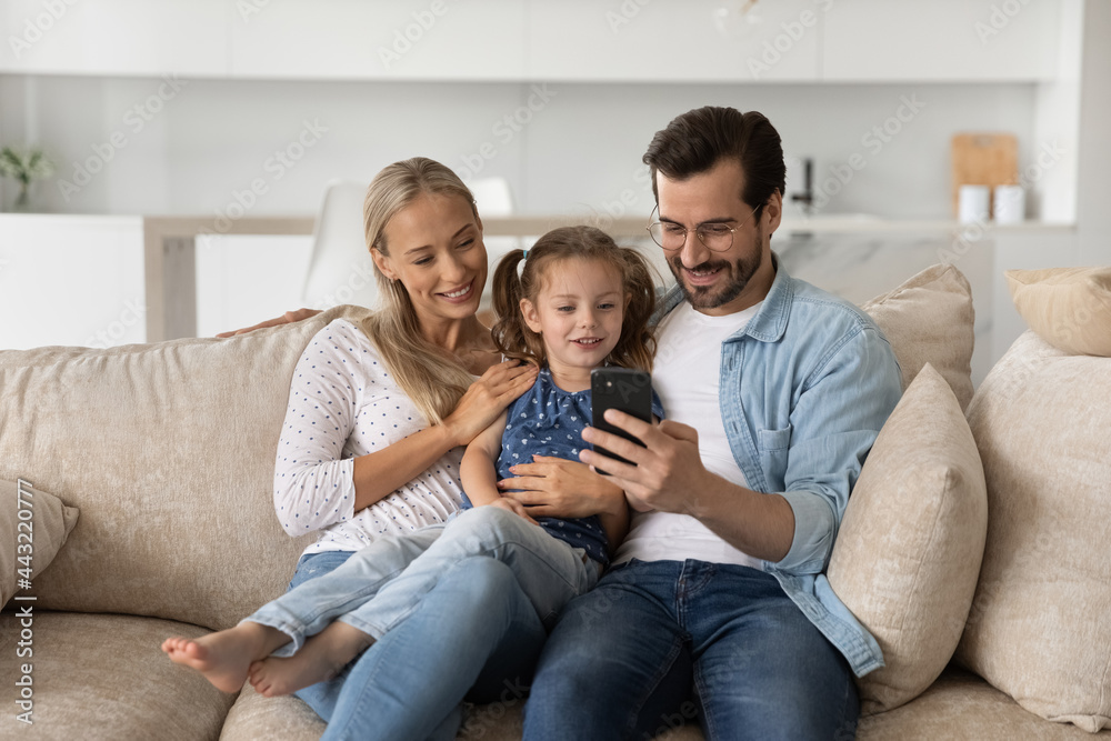 Happy family with little daughter using smartphone sitting on couch, smiling mother and father hugging adorable preschool child girl, looking at phone screen, watching video, having fun with device