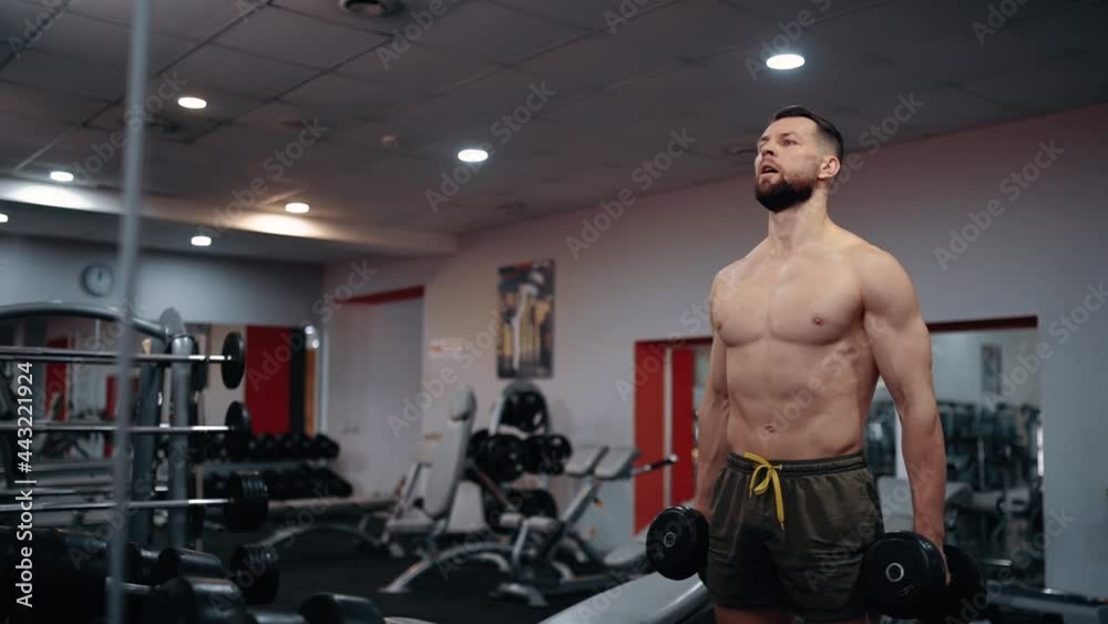 The muscular young man, with a bare chest and dumbbells in his hands, looks forward focused, tensing his muscles. Intensive training. Intense masculine energy. Healthy lifestyle. Sport concept
