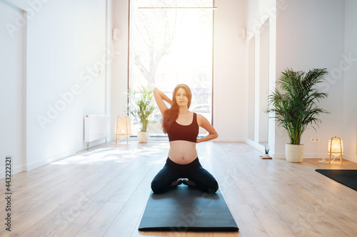 Sunlit image of a pregnant woman sitting on a mat, practicing yoga alone