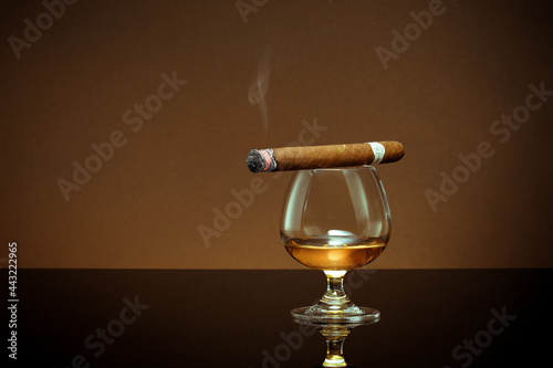 close up view of glass of cognac with cigar on top on color back. 