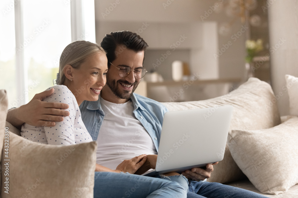 Close up happy young couple hugging using laptop together, relaxing sitting on couch at home, smiling wife and husband in glasses looking at computer screen, shopping or chatting in social network Close up happy young couple hugging using laptop together, relaxing sitting on couch at home, smiling wife and husband in glasses looking at computer screen, shopping or chatting in social network