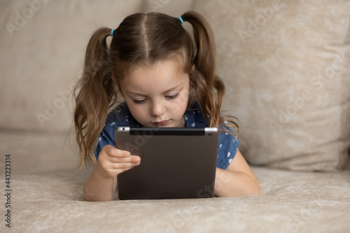 Close up curious little 7s girl using tablet lying on comfortable couch at home, pretty preschool child looking at device screen, playing game, studying online, spending leisure time with gadget photo