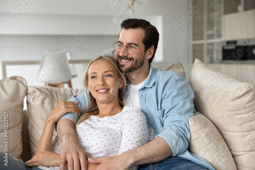 Dreamy happy young couple hugging, relaxing on couch together, smiling woman and man in glasses looking to aside in distance, visualizing good future planning, enjoying lazy leisure time on sofa