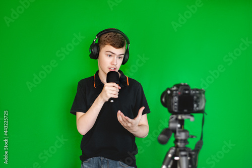 A boy teen blogger shoots a video on camera against a green background. A teenage Caucasian in a black T-shirt emotionally tells something for a blog.