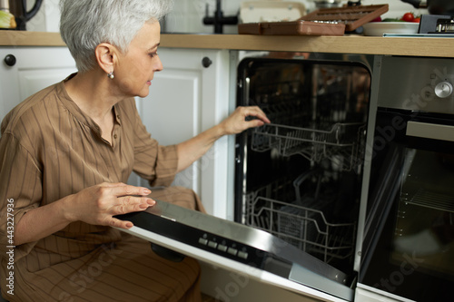 Serious woman posing at dishwasher in home kitchen. Middle-aged housewife using machine for washing dishes. Housekeeping, laundry and household concept