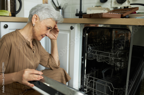 Upset middle-aged housewife sitting at broken dishwasher. Stressed woman looking at machine for washing dishes with sadness. Routine housekeeping and household concept