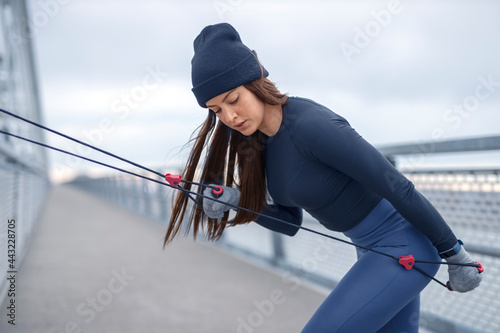 Young fitness woman in winter blue sportswear doing biceps exercises with resistance band cord on bridge during cold day