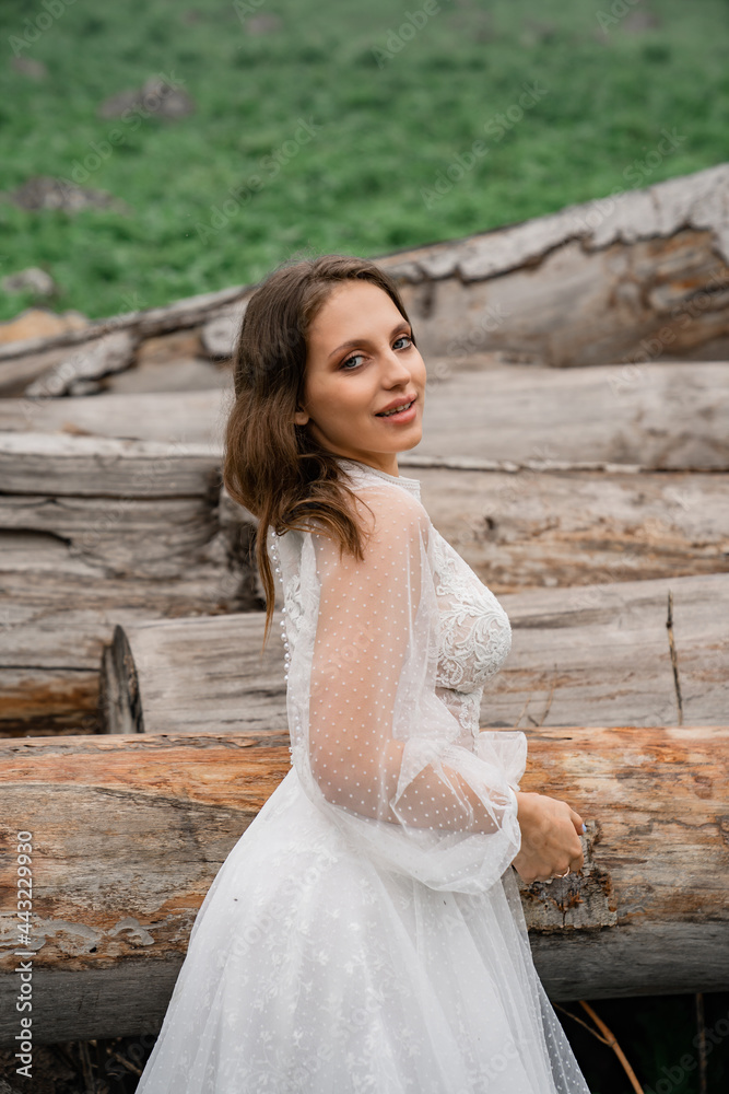 The bride in a white wedding dress stands in a field by the logs.