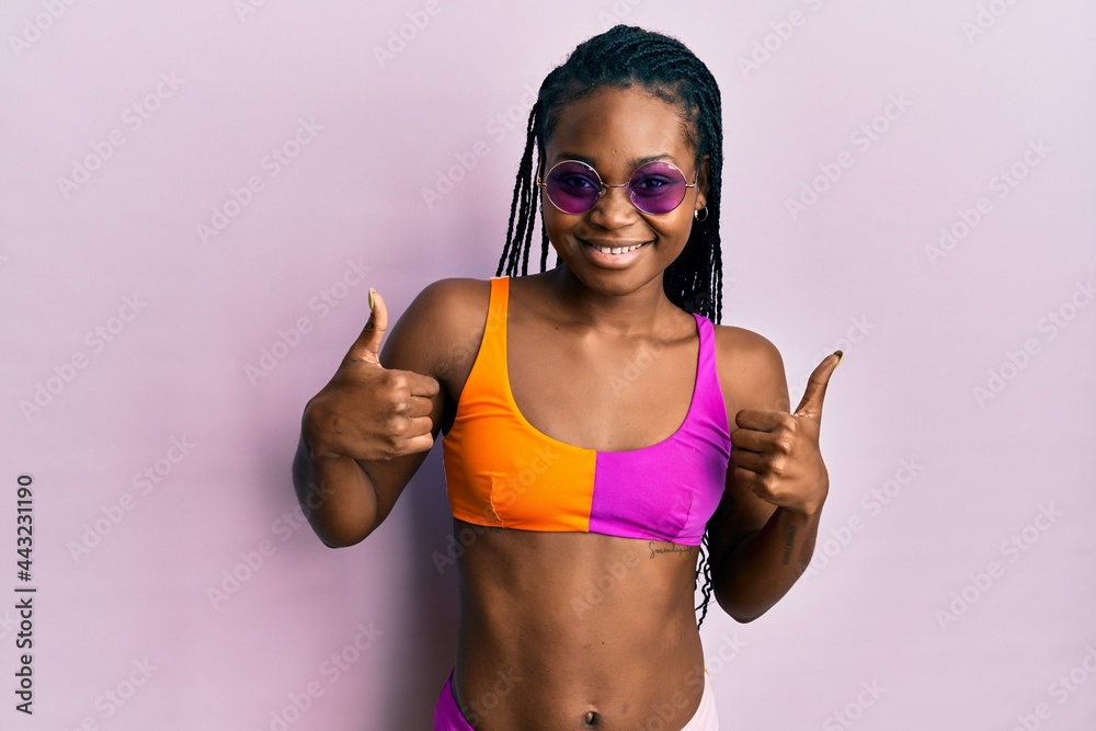 Young african american woman wearing bikini and sunglasses success sign doing positive gesture with hand, thumbs up smiling and happy. cheerful expression and winner gesture.