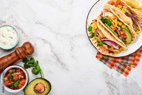 Mexican tacos with grilled chicken, avocado, corn kernels, tomato, onion, cilantro and salsa at white stone table. Traditional Mexican and Latin american street food. Top view.