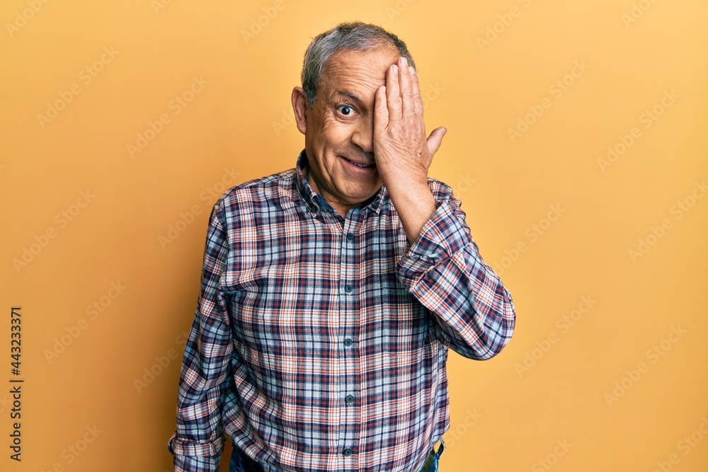 Handsome senior man with grey hair wearing casual shirt covering one eye with hand, confident smile on face and surprise emotion.