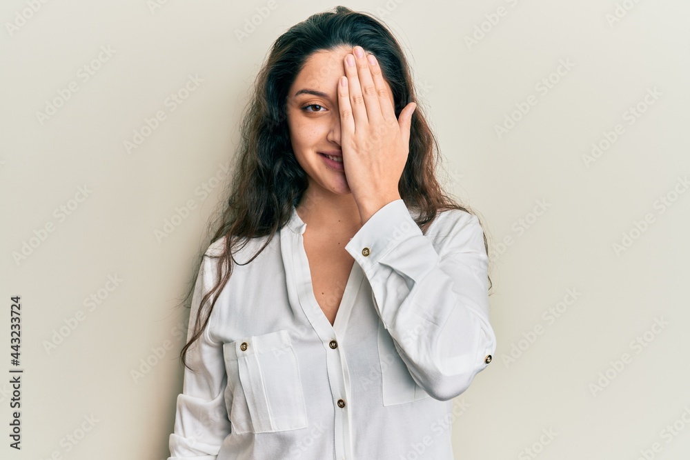 Beautiful middle eastern woman wearing casual clothes covering one eye with hand, confident smile on face and surprise emotion. Beautiful middle eastern woman wearing casual clothes covering one eye with hand, confident smile on face and surprise emotion.