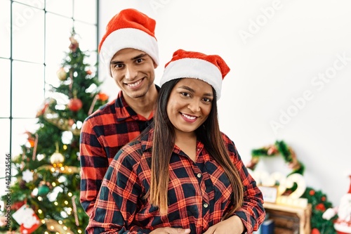 Young latin couple smiling happy and hugging standing by christmas tree at home.
