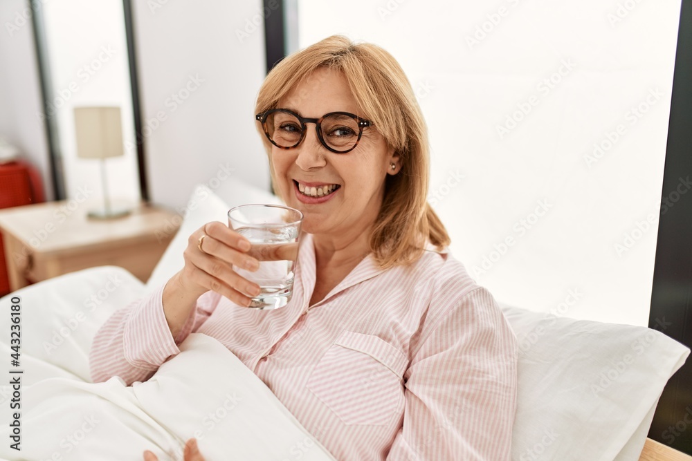 Middle age blonde woman smiling happy drinking glass of water lying on the bed at home.