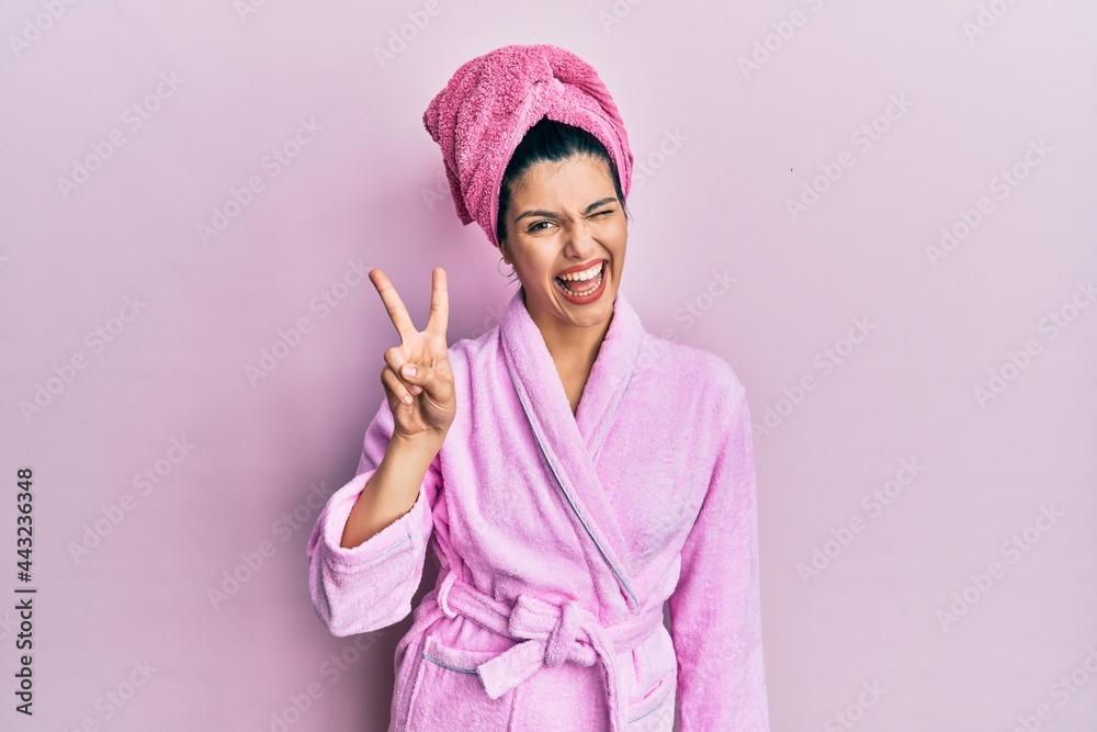 Young hispanic woman wearing shower towel cap and bathrobe smiling with happy face winking at the camera doing victory sign. number two. - obrazy, fototapety, plakaty 