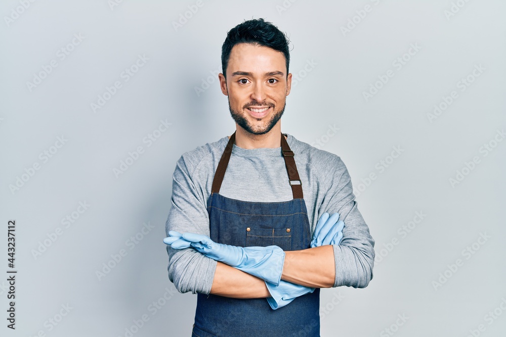 Young hispanic man wearing apron and gloves happy face smiling with crossed arms looking at the camera. positive person. Young hispanic man wearing apron and gloves happy face smiling with crossed arms looking at the camera. positive person.