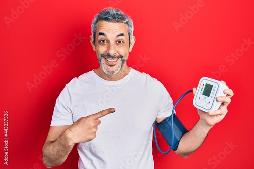 Handsome middle age man with grey hair using blood pressure monitor smiling happy pointing with hand and finger photo