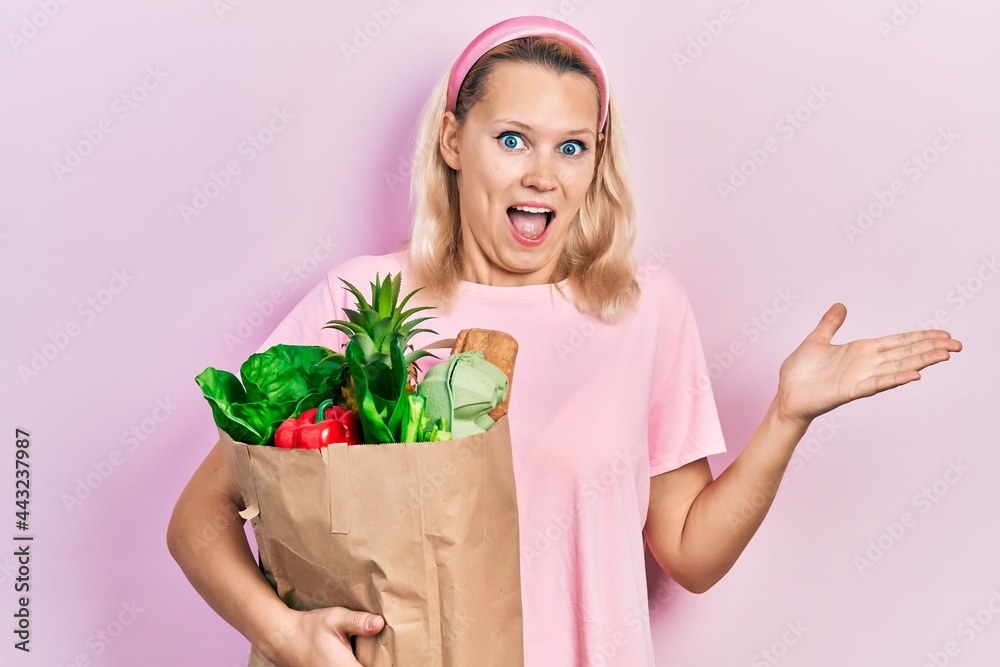 Beautiful caucasian blonde woman holding paper bag with groceries celebrating achievement with happy smile and winner expression with raised hand Beautiful caucasian blonde woman holding paper bag with groceries celebrating achievement with happy smile and winner expression with raised hand
