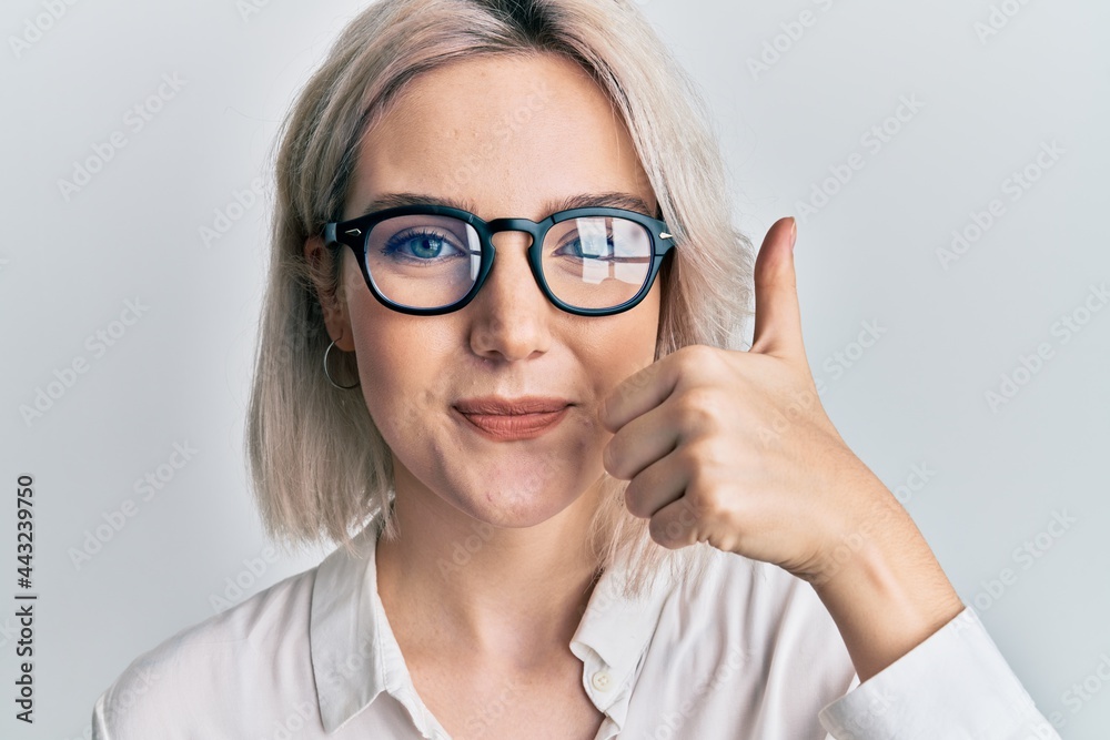 Young blonde girl wearing casual clothes and glasses smiling happy and positive, thumb up doing excellent and approval sign Young blonde girl wearing casual clothes and glasses smiling happy and positive, thumb up doing excellent and approval sign