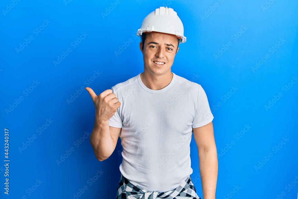 Handsome young man wearing builder uniform and hardhat smiling with happy face looking and pointing to the side with thumb up.