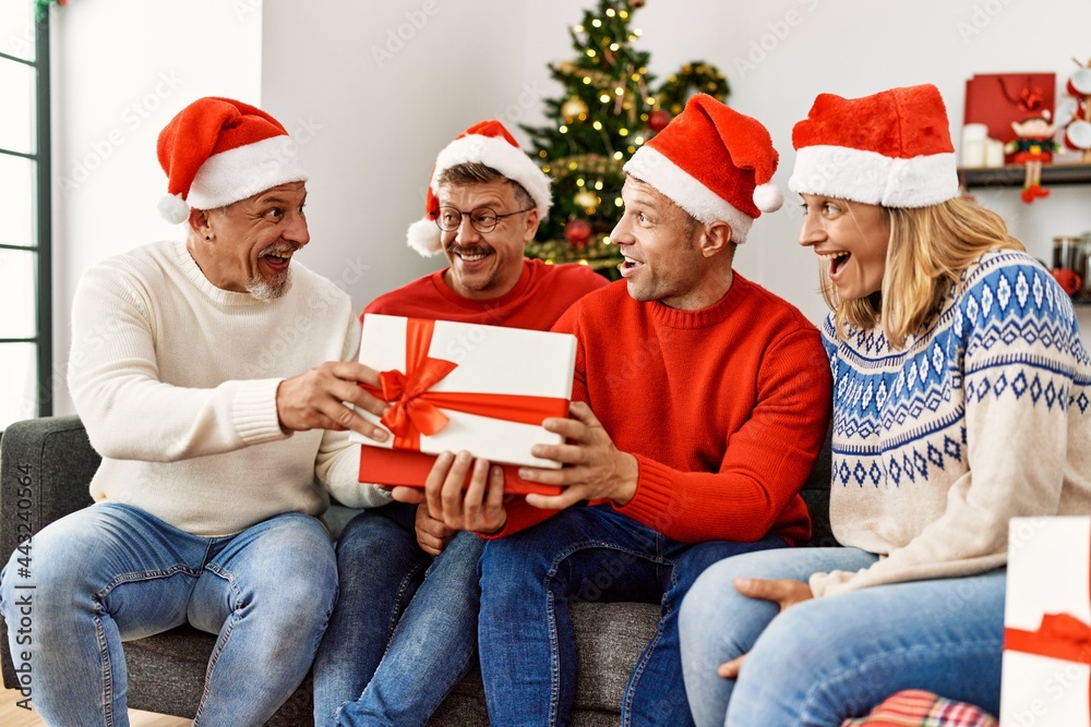 Fototapeta premium Group of middle age friends holding gifts sitting on the sofa by christmas tree at home.
