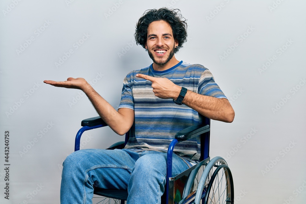 Handsome hispanic man sitting on wheelchair amazed and smiling to the camera while presenting with hand and pointing with finger. Handsome hispanic man sitting on wheelchair amazed and smiling to the camera while presenting with hand and pointing with finger.
