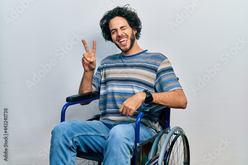 Handsome hispanic man sitting on wheelchair smiling with happy face winking at the camera doing victory sign with fingers. number two.