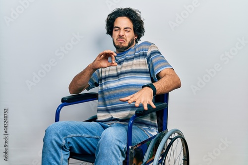 Handsome hispanic man sitting on wheelchair disgusted expression, displeased and fearful doing disgust face because aversion reaction.