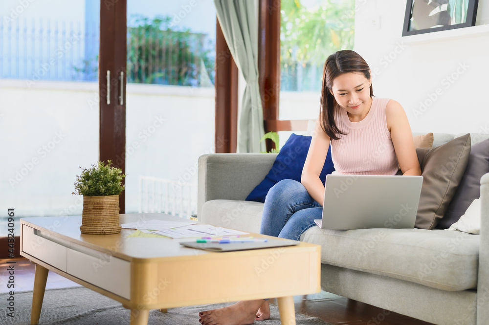Happy beautiful Asian woman using laptop working at home on sofa in living room Happy beautiful Asian woman using laptop working at home on sofa in living room