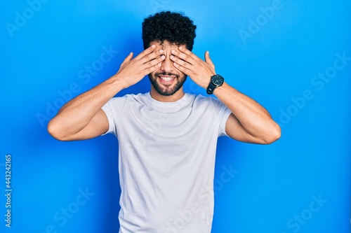 Young arab man with beard wearing casual white t shirt covering eyes with hands smiling cheerful and funny. blind concept.