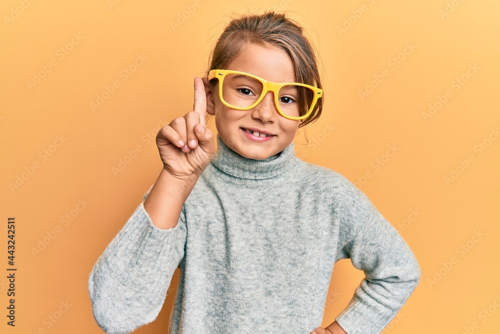 Little beautiful girl wearing casual clothes and yellow glasses smiling with an idea or question pointing finger with happy face, number one