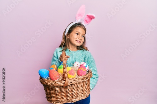 Little beautiful girl wearing cute easter bunny ears holding wicker basket with colored eggs smiling looking to the side and staring away thinking.