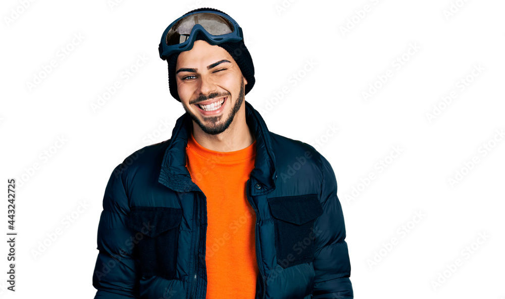 Young hispanic man with beard wearing snow wear and sky glasses winking looking at the camera with sexy expression, cheerful and happy face.