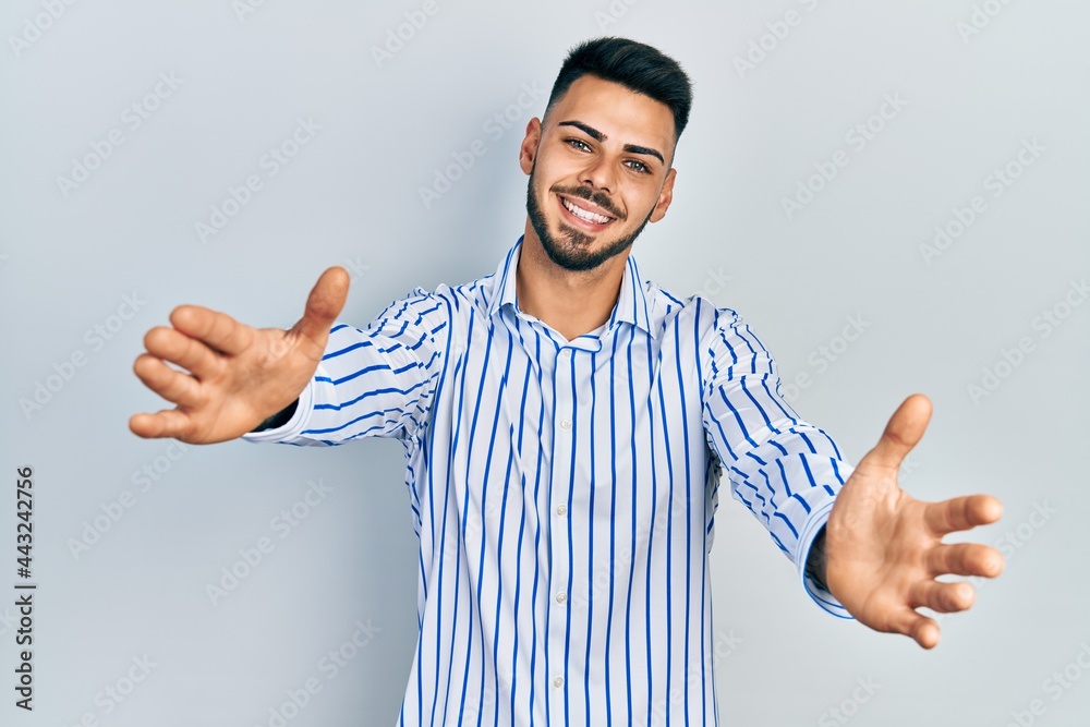 Young hispanic man with beard wearing casual striped shirt looking at the camera smiling with open arms for hug. cheerful expression embracing happiness. Young hispanic man with beard wearing casual striped shirt looking at the camera smiling with open arms for hug. cheerful expression embracing happiness.
