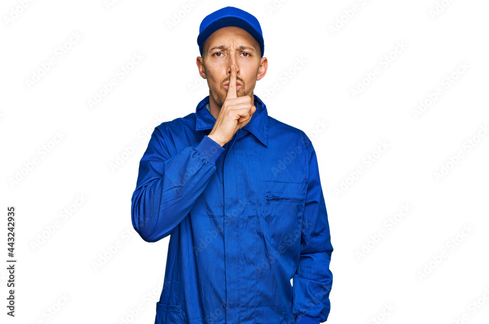 Bald man with beard wearing builder jumpsuit uniform asking to be quiet with finger on lips. silence and secret concept.