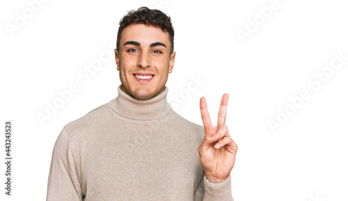 Hispanic young man wearing casual turtleneck sweater smiling with happy face winking at the camera doing victory sign. number two.