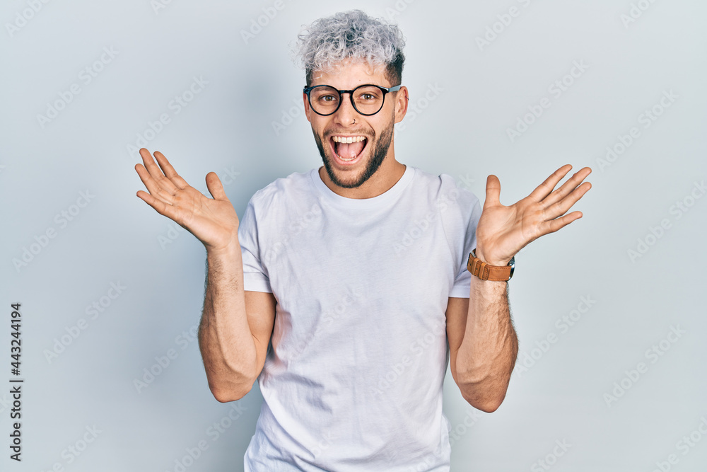 Naklejka premium Young hispanic man with modern dyed hair wearing white t shirt and glasses celebrating crazy and amazed for success with arms raised and open eyes screaming excited. winner concept