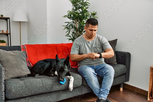 Young latin man and dog sitting on the sofa at home checking the time on wrist watch, relaxed and confident © Krakenimages.com