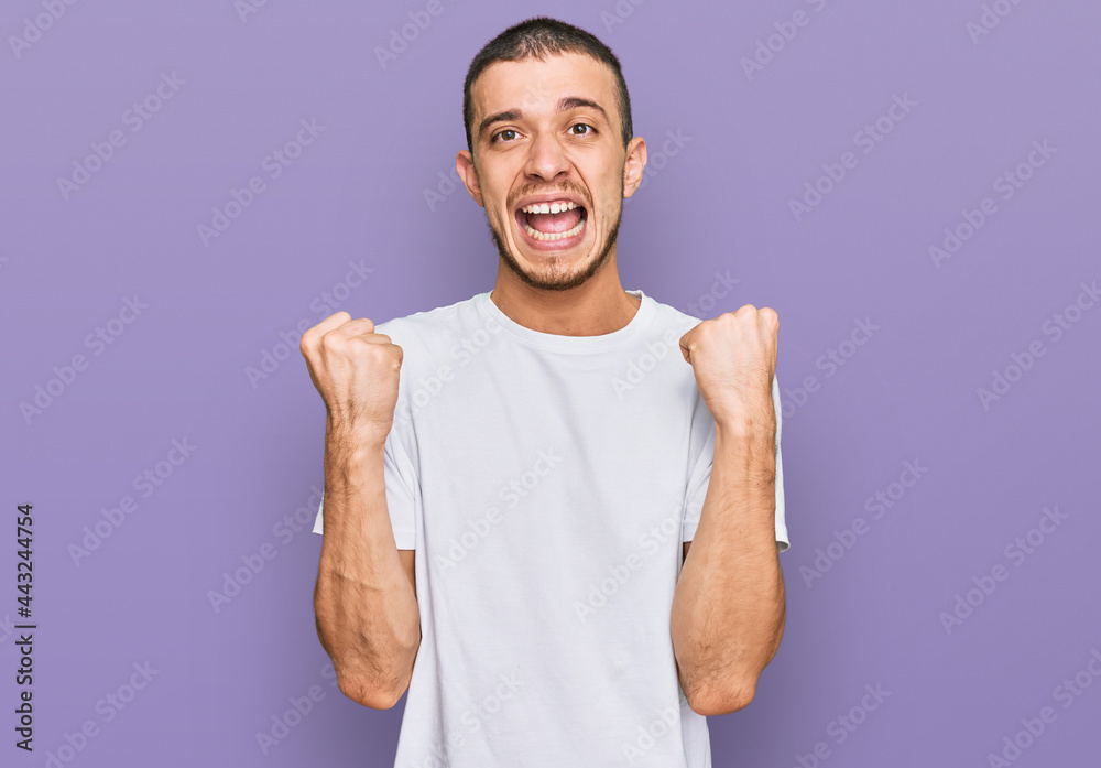 Hispanic young man wearing casual white t shirt celebrating surprised and amazed for success with arms raised and open eyes. winner concept. Hispanic young man wearing casual white t shirt celebrating surprised and amazed for success with arms raised and open eyes. winner concept.