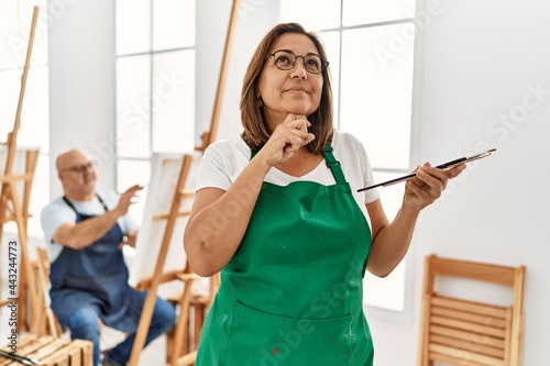 Middle age hispanic woman at art studio serious face thinking about question with hand on chin, thoughtful about confusing idea