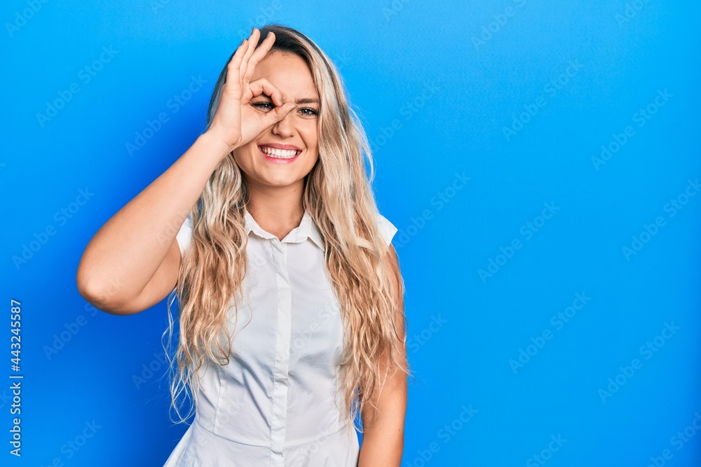 Beautiful young blonde woman wearing casual white shirt doing ok gesture with hand smiling, eye looking through fingers with happy face.