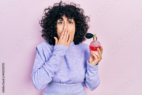 Young middle east woman holding perfume covering mouth with hand, shocked and afraid for mistake. surprised expression