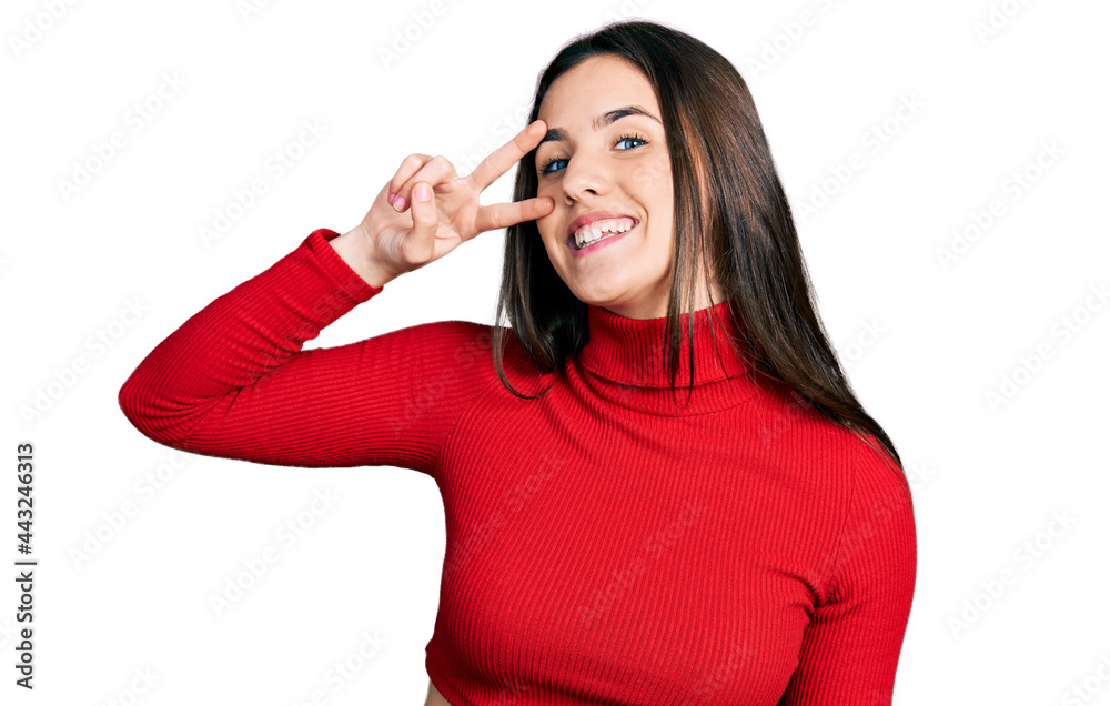 Young brunette teenager wearing red turtleneck sweater doing peace symbol with fingers over face, smiling cheerful showing victory